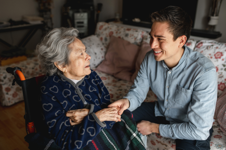 Young man sitting next to an elder woman in wheelchair taking her hands while talking and smiling